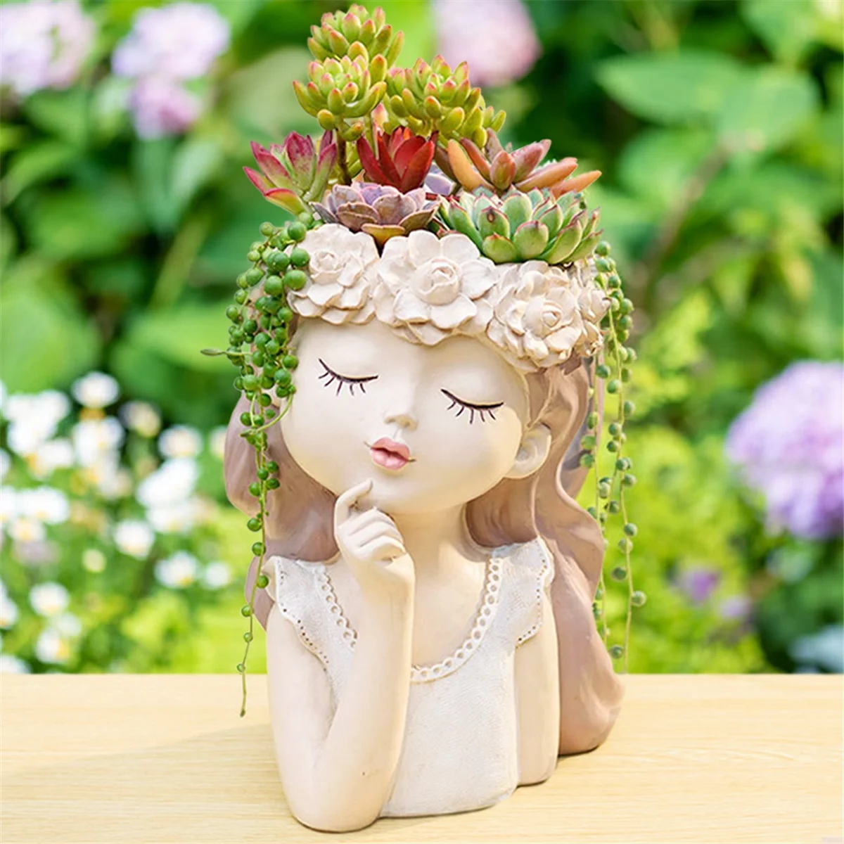 A whimsical planter shaped like a girl’s face with closed eyes and a serene expression, adorned with succulents and trailing greenery as her "hair." The planter is set on a wooden surface with a garden backdrop.